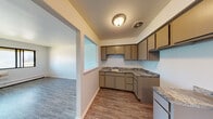 This single-floor unit offers a straightforward, functional layout connecting the kitchen, living area, and additional rooms via a cohesive open floor plan. Wood-look vinyl flooring runs consistently throughout the space, providing a clean, uniform appearance that ties each area together.

The kitchen is well-equipped with an L-shaped configuration featuring taupe cabinetry extending to the upper walls, offering generous storage capacity. Stone-patterned laminate countertops provide ample prep space, complemented by a stainless steel sink. A pass-through window opens directly to the living area, enhancing the sense of openness and connectivity between the two spaces.

The living area is spacious and bright, with white walls and a neutral palette that create a versatile, move-in-ready environment. Natural light enters through sliding and large fixed windows in the various rooms, some of which offer views of the surrounding neighborhood. Baseboard heating units are present throughout, along with a wall-mounted air conditioning unit in one room, ensuring year-round climate control.

The bathroom features a white vanity with an integrated sink, a wall-mounted mirror with overhead lighting, and matching wood-look flooring. The overall ambiance of the unit is clean, practical, and well-maintained.