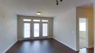 This single-floor space offers a clean, modern aesthetic defined by rich, dark hardwood flooring that extends throughout the main living area. A set of French doors framed by tall sidelights and transom windows above serves as a prominent architectural feature, allowing natural light to flood the interior and creating a strong visual connection to the outdoors. The white-trimmed windows are fitted with horizontal blinds, offering flexible light control while maintaining a polished look. The walls are finished in a neutral, warm beige tone that complements the dark flooring and contributes to an open, airy feel. Ceiling lighting includes both a classic flush-mount fixture and a modern adjustable track light system, providing versatile illumination options suitable for a range of uses. White baseboards add a crisp, finished detail throughout the room. The overall layout is open and unobstructed, maximizing usable floor space and offering flexibility in furniture arrangement or function. The combination of natural light, quality flooring, and thoughtful lighting fixtures gives this space a balanced, well-appointed character that is both functional and visually appealing.