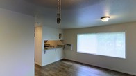 This single-floor space features an open, efficient layout that combines the living area and kitchen into one cohesive environment. The living area is finished with durable hardwood-style flooring that extends throughout, adding warmth to the neutral-toned walls. A large window fitted with blinds allows natural light to fill the room while also housing a wall-mounted air conditioning unit for climate control. A pull-chain light fixture is centrally positioned in the living space. The kitchen is accessible via a pass-through opening in the dividing wall, complete with a fold-down countertop supported by metal brackets — a practical feature that can serve as a breakfast bar or additional prep surface. The kitchen area includes an electric stovetop and features tile backsplash detailing. The textured ceiling adds subtle visual character throughout the space. Overall, the unit presents a straightforward, functional design with an emphasis on maximizing usable space within a compact footprint, making it well-suited for a single occupant or small household.