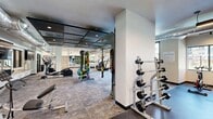 This modern fitness center offers a spacious, single-floor layout thoughtfully designed to accommodate a wide range of workout disciplines. The space is divided into distinct functional zones, each purpose-built for different training needs. The cardio area features multiple treadmills and stationary bikes positioned along a wall of large floor-to-ceiling windows, filling the space with natural light and providing outdoor views — creating an open, energizing atmosphere during workouts.

The strength training zone is anchored by a full cable machine system and a well-stocked dumbbell rack displayed along mirrored walls, which visually expand the space and assist with form monitoring. A dedicated functional fitness area includes TRX suspension trainers, medicine balls, and kettlebells neatly organized on tiered storage racks. Additional equipment such as rowing machines and a stair climber are distributed throughout the floor.

Exposed industrial-style ductwork runs along the ceiling alongside sleek linear LED lighting and recessed fixtures, lending a contemporary aesthetic to the space. The flooring transitions between durable rubber matting in high-impact zones and stone-textured tile in open areas. Built-in cabinetry and storage solutions are integrated along one wall, contributing to an organized, professional environment. The overall design balances functionality with a modern, clean aesthetic suitable for serious fitness use.