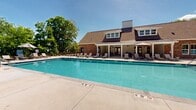 Located in Murfreesboro, Tennessee, this community amenity space centers around a generously sized outdoor swimming pool that serves as the focal point of the outdoor recreation area. The pool is enclosed by a sleek black iron fence and flanked by a wide, open concrete sun deck furnished with lounge chairs and patio umbrellas, providing ample space for relaxation and sunbathing under the Tennessee sky.

The adjacent clubhouse is a charming structure featuring classic brick exterior, a pitched shingle roof with dormer windows, and a covered rear porch outfitted with ceiling fans and outdoor dining furniture — ideal for shaded gatherings. The covered porch opens directly toward the pool area, creating a seamless indoor-outdoor connection.

A separate outdoor seating area with a circular fire pit arrangement and additional lounge chairs adds another social gathering spot within the fenced complex. Mature trees line the perimeter, offering natural privacy and a lush, green backdrop throughout the warmer months. The overall setting is well-maintained and designed for community enjoyment, combining practical amenities with an inviting, resort-style atmosphere in a residential neighborhood setting.