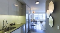This single-floor studio is designed to maximize efficiency without sacrificing style. The open-plan layout seamlessly integrates the kitchen, dining, and living/sleeping areas into one cohesive space. The kitchen runs along one wall and is outfitted with high-gloss, floor-to-ceiling cabinetry in a cool grey-green tone, providing ample storage. Stainless steel appliances, including a full-sized refrigerator and dishwasher, lend a contemporary, polished look. Under-cabinet lighting illuminates the countertops, enhancing functionality. A starburst pendant light fixture serves as a striking focal point, adding a sculptural, artistic element to the space. The dining area accommodates a small round table with modern chairs, positioned to take advantage of the generous natural light streaming through a wide band of windows spanning the far wall. The living and sleeping area benefits from this same bright exposure, creating an airy, open ambiance. Polished concrete floors run throughout, contributing to the space's modern industrial aesthetic. The overall design is clean, minimal, and cohesive, making thoughtful use of every available area.