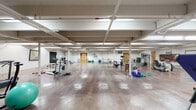 This spacious single-floor fitness and recreation facility offers a generous open layout designed to accommodate a wide variety of physical activities. The space is characterized by polished concrete floors that reflect overhead fluorescent lighting, creating a bright and functional atmosphere throughout. Exposed structural beams and support columns run across the ceiling, lending the space an industrial aesthetic that complements its active-use purpose.

The main workout area is generously proportioned, providing ample room for both equipment-based and open floor training. Cardio machines, including stationary bikes, ellipticals, and a treadmill, are positioned throughout the space, alongside free weight stations and a weight bench. Stability and flexibility training is well supported with stability balls, BOSU trainers, kettlebells, resistance equipment, inversion tables, and yoga mats.

A dedicated recreation zone features a full-size ping-pong table and a wall-mounted scoreboard, offering a lively space for recreational activities. Motivational wall art and artwork pieces are displayed throughout, adding personality and energy to the environment. Multiple wooden doors provide access to additional rooms and utility areas. A whiteboard and informational panels along the walls support organized programming. The overall ambiance is functional, active, and well-suited for fitness, rehabilitation, or multipurpose recreational use.