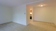 This single-floor space offers a straightforward and functional layout designed for comfortable everyday living. The main living area features wall-to-wall carpeting in a neutral beige tone, creating a warm and understated foundation throughout the space. Smooth, light-colored walls contribute to a clean, bright ambiance, while a flush-mount ceiling fixture provides consistent overhead lighting.

An open passageway connects the living area to the kitchen, which is efficiently arranged and well-equipped. The kitchen includes warm wood-toned upper cabinetry, a white electric range with a black oven, and additional built-in appliances. The kitchen flooring transitions to a lighter surface, visually distinguishing it from the carpeted living space.

The overall design is simple and practical, with a neutral color palette throughout that creates a cohesive, move-in-ready feel. The open sightline between the living area and kitchen adds a sense of connectivity, making the space feel accessible and easy to navigate. This unit is well-suited for those seeking a tidy, no-frills living environment with all essential amenities close at hand.