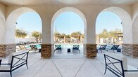 Located at 6710 W Doe Ave in Visalia, California, this outdoor amenity facility at Tuscan Village showcases a Mediterranean-inspired design that defines the entire space. At its centerpiece sits a large, rectangular swimming pool surrounded by a generously sized concrete pool deck lined with modern chaise lounges, providing ample room for sunbathing and relaxation under the Central Valley sun.

The covered patio structure is defined by elegant stucco arches resting on stacked-stone columns, offering a shaded retreat with ceiling fans and comfortable seating overlooking the pool. This architectural element establishes a refined, resort-like ambiance throughout the space.

On the opposite side of the pool, a series of wooden pergolas supported by natural stacked-stone pillars provide additional shaded lounging areas outfitted with cushioned outdoor furniture. The pergolas frame views of mature olive and ornamental trees that add greenery and character to the enclosed grounds.

A dedicated BBQ and dining pavilion with a metal canopy roof offers an outdoor cooking and gathering area with table seating, ideal for social gatherings. The perimeter of the space features curated landscaping with flowering plants and shrubs set against clean white privacy walls, creating a polished, enclosed environment. Restroom facilities and pool safety signage are also present, supporting the functional needs of the space.