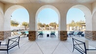 Located at 6710 W Doe Ave in Visalia, California, this outdoor amenity facility at Tuscan Village showcases a Mediterranean-inspired design that defines the entire space. At its centerpiece sits a large, rectangular swimming pool surrounded by a generously sized concrete pool deck lined with modern chaise lounges, providing ample room for sunbathing and relaxation under the Central Valley sun.

The covered patio structure is defined by elegant stucco arches resting on stacked-stone columns, offering a shaded retreat with ceiling fans and comfortable seating overlooking the pool. This architectural element establishes a refined, resort-like ambiance throughout the space.

On the opposite side of the pool, a series of wooden pergolas supported by natural stacked-stone pillars provide additional shaded lounging areas outfitted with cushioned outdoor furniture. The pergolas frame views of mature olive and ornamental trees that add greenery and character to the enclosed grounds.

A dedicated BBQ and dining pavilion with a metal canopy roof offers an outdoor cooking and gathering area with table seating, ideal for social gatherings. The perimeter of the space features curated landscaping with flowering plants and shrubs set against clean white privacy walls, creating a polished, enclosed environment. Restroom facilities and pool safety signage are also present, supporting the functional needs of the space.