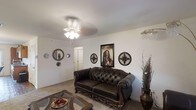 This single-floor space offers a welcoming and character-rich interior with a distinctly eclectic aesthetic. The living area serves as the focal point of the space, featuring an ornately carved dark leather tufted sofa that anchors the room with a sense of classic elegance. A glass-top coffee table complements the seating arrangement, adding a touch of refinement to the space.

The walls are adorned with a curated collection of decorative pieces, including ornamental round mirrors and cross medallions, alongside a prominent framed Bob Marley "Soul Rebel" poster that lends the space a bold, personalized character. A tall decorative vase with dried botanical accents adds an organic, artistic element to the corner of the room.

The flooring is carpeted, contributing to a warm and comfortable atmosphere throughout. Overhead, a ceiling fan with an integrated light fixture provides both ventilation and ambient lighting, while a small chandelier is visible in the adjoining area, hinting at additional living or dining space. A white interior door leads to further rooms within the layout.

Overall, the space presents a lived-in, expressive environment that blends ornate furniture with eclectic décor, creating a distinctive and personal atmosphere throughout.