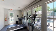 This fitness center is a single-story facility designed to support a well-rounded workout routine. The space is outfitted with a variety of cardio equipment, including treadmills, an elliptical trainer, a stair climber, and a rowing machine, all positioned to take advantage of the room's generous natural light. Multiple large double-hung windows line the walls, offering views of the adjacent outdoor pool area and lush tree canopy, creating an open and energizing atmosphere during workouts.

The flooring throughout is durable rubberized material, practical for both cardio and strength training activities. A multi-station cable weight machine provides comprehensive strength training options and is situated in a dedicated area with ample surrounding space. Recessed lighting in the textured ceiling supplements the natural light, ensuring the space remains well-lit at all times.

The walls are painted in a neutral gray tone, and decorative artwork—including a colorful bicycle-themed canvas and abstract pieces—adds visual interest throughout the space. Multiple entry and exit points, including a glass-paneled door leading to an outdoor deck area, enhance accessibility and ventilation. Overall, the fitness center presents a bright, functional, and well-maintained environment suited for a variety of fitness activities.