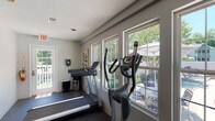 This fitness center is a single-story facility designed to support a well-rounded workout routine. The space is outfitted with a variety of cardio equipment, including treadmills, an elliptical trainer, a stair climber, and a rowing machine, all positioned to take advantage of the room's generous natural light. Multiple large double-hung windows line the walls, offering views of the adjacent outdoor pool area and lush tree canopy, creating an open and energizing atmosphere during workouts.

The flooring throughout is durable rubberized material, practical for both cardio and strength training activities. A multi-station cable weight machine provides comprehensive strength training options and is situated in a dedicated area with ample surrounding space. Recessed lighting in the textured ceiling supplements the natural light, ensuring the space remains well-lit at all times.

The walls are painted in a neutral gray tone, and decorative artwork—including a colorful bicycle-themed canvas and abstract pieces—adds visual interest throughout the space. Multiple entry and exit points, including a glass-paneled door leading to an outdoor deck area, enhance accessibility and ventilation. Overall, the fitness center presents a bright, functional, and well-maintained environment suited for a variety of fitness activities.