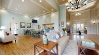 Located at 1500 Royal Gold Dr in Columbus, Ohio, this single-story community clubhouse offers a thoughtfully designed gathering space that balances comfort and functionality. The grand entrance greets visitors with soaring ceilings, warm hardwood-style flooring, and an elegant rotunda-style foyer accented by large windows that flood the space with natural light and views of the surrounding greenery.

The expansive main gathering area features a soft, spa-like palette of sage green and neutral tones, anchored by a stone accent wall with a built-in beverage station complete with granite countertops, white cabinetry, and a mosaic tile backsplash. A wall-mounted television, chandelier lighting, and coffered ceiling details add refinement to the open layout.

The dining area accommodates multiple large farmhouse-style wooden tables paired with teal accent chairs, ideal for community events and group gatherings. Adjacent seating nooks with oversized windows overlook the outdoor pool area, providing bright, airy spots for relaxed conversation.

A private office space features textured wall coverings, a modern workstation, and comfortable seating for guests, offering a quiet and professional environment. Mirrored sliding doors elegantly separate flexible spaces while maintaining an open visual flow. Throughout, the design balances welcoming warmth with polished, community-focused functionality.