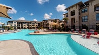 Located at 1930 Kingsley Dr in Pearland, Texas, this outdoor amenity space showcases a stunning resort-style pool complex designed for relaxation and recreation. The centerpiece is a large, freeform pool with crystal-clear blue water, gently curving edges, and a shallow wading area, all finished with a broad, light-toned paver surround that provides ample space for sunbathing and socializing. Multiple lounge chairs, colorful accent seating, and shade umbrellas are thoughtfully arranged around the deck, offering both sun-drenched and shaded retreat options. A separate spa or splash feature is nestled within the pool area, adding an additional layer of leisure amenity. The surrounding multi-story buildings are constructed with classic stone and stucco facades, creating an upscale architectural backdrop. Lush tropical landscaping, including mature palm trees, ornamental shrubs, and layered greenery, frames the space and provides a vibrant, resort-like atmosphere. Wrought iron fencing encloses the area, ensuring privacy and security while maintaining open sightlines to the beautifully maintained grounds. The outdoor space benefits from abundant natural Texas sunlight, making it an inviting destination throughout the day.