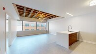 This single-floor unit blends industrial and contemporary design elements throughout its open layout. The living and kitchen area immediately draws attention with its striking exposed waffle-slab concrete ceiling, partially framed by a smooth white drywall soffit. A sculptural sputnik chandelier anchors the space, complementing the clean, modern aesthetic. Large floor-to-ceiling windows span the exterior wall, flooding the interior with natural light and offering urban views of the surrounding cityscape.

The kitchen is defined by a central island with a white countertop and integrated sink, paired with warm wood cabinetry that extends to a dedicated cooking wall. Stainless steel appliances, including a refrigerator, range, and microwave, are set against a teal mosaic tile backsplash that adds a vibrant accent. Light-toned flooring runs throughout the main living and kitchen zones, contributing to a bright, open feel.

The bedroom is a calm, neutral-toned retreat finished with plush carpet, a standard closet, and a window admitting natural light. Additional rooms maintain the same soft, understated palette with carpet flooring and clean white walls. The bathroom features a streamlined wood vanity with a white countertop and a walk-in shower clad in bold teal subway tile, providing a cohesive design detail that ties back to the kitchen's accent elements.