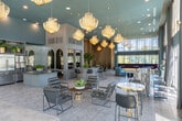 Located at 100 Zoo Lane in Santa Ana, California, this stylish communal lounge at Parks on Muir is a single-floor space that exudes contemporary elegance across a generously proportioned open floor plan. The interior immediately impresses with soaring double-height ceilings adorned with a striking array of sculptural gold orb pendant lights that cast a warm, ambient glow throughout. Floor-to-ceiling windows wrapped in layered sheer and mocha drapery flood the space with natural light while framing urban views of the surrounding Santa Ana skyline.

The main lounge area features a carefully curated seating arrangement, including a curved deep-blue velvet sofa, plum-toned banquette seating, and sleek black leather chairs — all set atop a bold geometric-patterned tile floor. Sculptural art pieces and lush tropical greenery add organic contrast to the refined palette of teal, grey, and gold. A fully appointed kitchen island area showcases rich marble-look countertops, mirrored mosaic tile backsplash, arched display niches, and stainless steel appliances, complemented by bar seating for casual gatherings. A flexible event area with dramatic floor-length gold drapery and curated wall art provides an intimate backdrop for private functions. Glass entry doors bearing the Parks on Muir branding complete the polished, welcoming atmosphere of this exceptional amenity space.