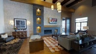 This single-story space showcases a compelling blend of modern and rustic design elements. The living area is anchored by a floor-to-ceiling stone accent wall, which serves as the room's focal point. Built into the wall are illuminated display niches framed in warm wood and slate-toned material, adding visual depth alongside a sleek linear fireplace below. A flat-screen television is mounted centrally within the composition, integrating media seamlessly into the architectural design.

The soaring ceiling features exposed dark wood beams that contrast beautifully with the light stone and hardwood flooring, creating a warm, lodge-inspired atmosphere. Pendant lighting with cylindrical shades hangs at varying heights, contributing to the layered illumination throughout the space.

The seating area is thoughtfully arranged around a geometric-patterned area rug in earth tones, complemented by neutral upholstered furniture and a substantial wood coffee table. A barn-style sliding door is visible in the background, enhancing the rustic-modern character. Large windows allow natural light to filter in, further brightening the inviting, open layout.