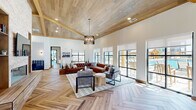 This well-appointed single-floor space offers a generous open layout designed for both gathering and relaxation. The interior is immediately distinguished by its soaring vaulted ceilings clad in warm wood planking, accented by exposed wood beams that lend a refined yet inviting character throughout. Herringbone-patterned hardwood flooring flows seamlessly across the entire space, adding a sophisticated visual texture underfoot.

The main living area is expansive and sun-drenched, with floor-to-ceiling black-framed windows and glass doors that invite abundant natural light and provide direct views of an outdoor pool area. A statement drum chandelier anchors the lounge seating arrangement, which features tufted leather sectionals and woven accent chairs centered on a patterned area rug.

A dramatic stone fireplace wall with built-in shelving and a mounted television serves as a focal point in an adjoining lounge zone, accompanied by a billiards table and intimate bistro-style seating nooks with plaid upholstered banquettes and navy velvet chairs — ideal for casual socializing.

The kitchen and dining area incorporates a large wood-topped island, pendant lantern lighting, shiplap wall detailing, and a built-in window bench with navy accent walls and open shelving, creating a warm and functional gathering space. Black-framed glass double doors connect distinct zones while maintaining an open, airy flow throughout.