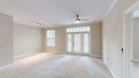 This single-story, 3-bedroom, 2-bathroom residence offers a thoughtfully designed floor plan that maximizes both comfort and functionality. The open living area is a standout feature, showcasing soft neutral tones, plush wall-to-wall carpeting, and crown molding that adds a refined architectural touch. A ceiling fan with light fixture keeps the space comfortable, while a set of French doors flanked by transom windows floods the room with natural light and provides direct access to an outdoor area. Track lighting adds additional illumination options. The kitchen is equipped with dark cabinetry and modern appliances, flowing seamlessly into the main living space as seen in the floor plan. The three bedrooms are finished in the same warm, neutral palette with plush carpeting throughout. The primary bedroom benefits from corner windows on two walls, offering abundant natural light and pleasant outdoor views of surrounding greenery. The secondary bedrooms are well-proportioned with natural light from individual windows. A dedicated laundry room houses a full-size front-loading washer and dryer set alongside a water heater, adding practical convenience. Clean white trim and baseboards throughout the residence contribute to a cohesive and polished aesthetic.