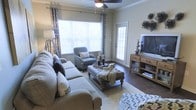 This single-floor residence offers a thoughtfully designed interior that balances comfort and style. The living room serves as the heart of the space, showcasing rich hardwood flooring that runs throughout and elegant crown molding that frames the room with a classic touch. Large windows dressed with patterned curtains flood the area with natural light, creating a warm and inviting atmosphere. A ceiling fan adds both function and character overhead. The room is generously proportioned, providing ample space for a full sectional sofa, an upholstered tufted ottoman that doubles as a coffee table, and a cozy accent chair. A wooden media console anchors one wall, offering open shelving and storage. Decorative wall art and layered textiles — including throw blankets and accent pillows — add depth and personality to the space. A glass-paneled exterior door allows additional light to pour in while providing access to an outdoor area, hinting at views and fresh air just beyond. The overall ambiance is relaxed yet refined, making this a comfortable and functional living environment suited for both everyday living and entertaining.