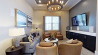 Located at 1680 Bluewater Way in Charleston, South Carolina, this single-floor space exudes a polished and welcoming ambiance through its thoughtful design and upscale finishes. The living area showcases a striking tray ceiling that adds architectural depth and dimension to the room, complemented by a grand circular chandelier with exposed Edison-style bulbs that serves as a bold focal point. Warm neutral tones across the walls and flooring create a cohesive, inviting atmosphere throughout. The seating arrangement is both stylish and functional, featuring a plush gray sofa accented with navy and yellow throw pillows, paired with curved caramel leather accent chairs that lend a contemporary yet cozy feel. A round marble-topped coffee table anchors the conversation area, while a sleek dark media console with cabinetry provides practical storage along the wall-mounted television side. Natural light filters in through a window dressed with layered valance and sheer drapery panels, brightening the space while maintaining a sense of warmth. Tasteful artwork adorns the walls, adding color and character. The overall design blends comfort and sophistication, making this Charleston space a refined and functional environment.