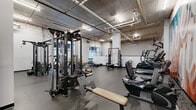 Located at 1 W Dickerson St in Dover, NJ, this fitness facility offers a comprehensive exercise environment spread across a single, generously sized floor. The space is defined by its industrial aesthetic, with exposed concrete ceilings, visible overhead piping, and structural columns that lend an urban, warehouse-style character throughout.

The exercise area is extensively equipped for both strength training and cardio. Strength equipment includes multi-cable functional trainers by Life Fitness, power racks with benches, and a variety of selectorized machines for targeted muscle training. The cardio section features a row of TRUE brand treadmills, ellipticals, and stair climbers. Full-length mirrors along one wall enhance the sense of space while supporting proper form during workouts. Rubber flooring throughout provides durability and safety underfoot.

A standout feature is a vibrant, large-scale motivational mural adorning one wall, adding energy and visual interest to the cardio area. Adjacent to the main gym floor, a dedicated indoor basketball court with a polished hardwood floor, regulation hoop, and padded wall panels offers space for court-based recreation and training.

Natural light filters in through windows along the perimeter, complemented by overhead fluorescent lighting. The overall ambiance is functional, energetic, and well-appointed, making this facility a versatile destination for a wide range of fitness activities.
