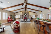 Located at 2857 S Lovers Ln in Visalia, California, this single-floor community clubhouse is defined by its dramatic vaulted ceilings adorned with rich, exposed wood beams that span the length of the main gathering space. The open-concept great room serves as the heart of the facility, anchored by a floor-to-ceiling stacked stone fireplace that draws the eye upward and creates a warm, inviting focal point. Hardwood-style flooring flows throughout, complementing the warm neutral tones of the walls and contributing to a cohesive, welcoming ambiance. Multiple seating areas are arranged throughout the great room, providing flexible space for socializing and relaxation. A full kitchen with rich wood cabinetry, stainless steel appliances, and a bar-height counter with seating offers excellent functionality for events and gatherings. A dedicated dining area with a large wooden table provides a more formal setting for meals. A separate game room features a full-size pool table, additional lounge seating, and a wall-mounted television, offering a casual retreat for entertainment. The formal entry foyer welcomes visitors with elegant tile flooring, crown molding, a chandelier, and graceful arched doorways. Plantation shutters and generous windows allow natural light to filter throughout the space, enhancing its bright and open character.