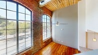This two-story loft at 1101 Hawthorne Lane in Charlotte, NC showcases a striking industrial-chic aesthetic with exposed original brick walls, warm hardwood floors, and distinctive wood-plank ceilings throughout. The main floor features soaring ceilings, large arched warehouse-style windows that flood the space with natural light, and an open-concept layout anchored by a granite-topped kitchen island with a stainless steel sink. A sleek staircase leads to the upper level, where two bedrooms offer wood ceilings and horizontal strip windows. The full bathroom features tile flooring, a granite vanity, and a tub-shower combo.