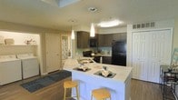 Located at 7301 W. Ohio Ave. in Lakewood, Colorado, this single-floor residence offers a thoughtfully designed open-concept layout that maximizes both function and comfort. The kitchen serves as a central highlight, featuring rich dark cabinetry paired with light countertops that create a striking contrast. A spacious peninsula with bar seating and elegant pendant lighting makes the kitchen ideal for casual dining and entertaining. Black appliances, including a full-size refrigerator and range, complement the modern aesthetic. Recessed and flush-mount ceiling fixtures provide ample illumination throughout. The kitchen flows seamlessly into the adjacent living areas, enhancing the open feel of the floor plan. Durable wood-style flooring runs throughout the main living space, offering both visual warmth and easy maintenance. Textured ceilings add a classic touch overhead. A hallway visible from the kitchen leads to additional rooms, including a bathroom with patterned curtain detail. Interior doors throughout are paneled in a clean, traditional style. The overall ambiance is modern yet inviting, with a neutral color palette and well-chosen finishes that lend the space a polished, move-in-ready appeal.