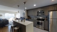 This single-floor space showcases a thoughtfully designed open-concept layout that integrates the kitchen, dining, and living areas into one cohesive environment. The kitchen is a standout feature, outfitted with sleek, dark wood-toned cabinetry that extends to the ceiling, maximizing storage capacity. A grey subway tile backsplash adds texture and visual interest, complementing the dark countertops throughout. Stainless steel appliances, including a freestanding range with a built-in overhead microwave, lend a polished, contemporary feel. A central island with a light-toned countertop provides additional prep space and doubles as a casual dining area, complete with industrial-style bar stools. Two modern pendant lights hang above the island, adding focused task lighting and a stylish design element. Recessed lighting throughout ensures the space remains bright and well-illuminated. The open sightlines extend into the living and dining areas, where large windows allow natural light to fill the space. Light tile flooring ties the kitchen zone together, while the overall design aesthetic maintains a clean, modern, and functional character throughout.