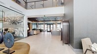 Located at 3001 S 21st Ave in Bozeman, Montana, this generously sized amenity space delivers a polished, contemporary experience across a single expansive floor. The grand lobby entrance features large-format light tile flooring, soaring double-height ceilings, and floor-to-ceiling glass entry doors that flood the space with natural light. An open-railed upper mezzanine overlooks the main floor, reinforcing the sense of verticality and openness.

The lounge area is anchored by a brick-accented fireplace wall with a mounted display, plush seating, and stylish pendant lighting, creating a welcoming social hub. Adjacent to the lounge, a glass-enclosed co-working room offers a focused workspace with modern seating and a built-in desk. A communal kitchen and dining area features a substantial island with leather seating, pendant lights, navy cabinetry, and open shelving — ideal for gatherings and events.

A striking staircase with cable and metal railings, set against a slatted wood accent wall, leads to the mezzanine level. A dedicated golf simulator room rounds out the amenities, offering an interactive entertainment experience. Throughout, the design blends warm wood tones, textured wall finishes, and sophisticated furnishings to create a refined yet approachable atmosphere.