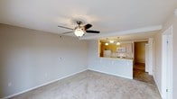 This single-floor residence offers a clean, open-concept layout designed for comfortable everyday living. The living area is generously sized, finished with soft neutral carpeting underfoot and crisp white walls that create a bright, airy atmosphere. A ceiling fan with a built-in light fixture anchors the room, providing both style and functional air circulation. The space transitions seamlessly into the kitchen through a pass-through breakfast bar, which features a white countertop with bar seating — ideal for casual dining or entertaining. The kitchen is well-appointed with warm wood-toned cabinetry, a white refrigerator, microwave, and range. Pendant lighting hangs above the kitchen area, adding a warm, inviting glow. The kitchen also features tile flooring, offering a practical contrast to the plush carpeting of the living space. The overall design is clean and functional, with a neutral color palette throughout that lends itself to a wide range of furnishing styles. The thoughtful open layout maximizes the sense of space, making the living and kitchen areas feel connected and cohesive. This well-maintained, move-in ready residence is an excellent choice for those seeking a comfortable and practical living environment.
