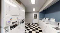 This shared laundry facility is designed for practical, everyday use and provides residents with a comprehensive set of amenities in a clean, organized environment. The space is finished with a distinctive black-and-white checkered tile floor that runs throughout, lending a classic aesthetic to the room. Walls are painted in neutral gray tones, with a bold blue accent wall highlighting the washer area, adding a visual distinction between functional zones.

The facility houses multiple top-load washing machines positioned along one wall within the blue-accented alcove, each equipped with card-operated payment terminals that accept major credit cards, offering a convenient cashless payment option. Alongside the washers, a row of stacked dryer units lines the opposite wall, also outfitted with card-reader payment systems. White beadboard wainscoting runs along the lower portion of the walls, contributing to the space's clean, finished appearance.

A dedicated folding area features a granite-topped counter surface along one wall, providing ample space for sorting and folding laundry. The facility is well-lit with recessed ceiling panels and is accessible via a glass-panel exterior door that allows natural light into the space. Overall, the facility is functional, well-maintained, and thoughtfully organized.