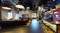 This single-floor space presents a striking industrial-chic aesthetic defined by exposed ceiling infrastructure, dark steel, concrete-textured walls, and warm wood accents. The expansive main lobby features large-format dark tile flooring, dramatic pendant lighting, and a set of glass-paneled entry doors that draw in natural light. A central lounge area anchors the space with a double-sided gas fireplace set into a concrete surround, surrounded by plush dark sofas and rich burgundy armchairs arranged on light hardwood platforms, creating an inviting contrast against the darker flooring. Adjacent to the lounge, a bar counter with industrial-style adjustable stools sits beneath pendant lights against a vivid red accent wall adorned with large-scale photography and graphic artwork. A staircase with steel railings leads to a mezzanine level with glass balustrades, further emphasizing the vertical scale of the interior. A separate dining or lounge alcove features warm amber-backlit shelving, red leather booth seating, and dark wood tables beneath cylindrical drum pendant lights. The elevator lobby provides a refined counterpoint, clad in natural oak wood paneling with a sculptural artwork niche at its center. Throughout, the layering of textures, curated art, and carefully controlled lighting creates a sophisticated, urban atmosphere.