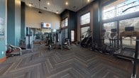 Located at 1121 3rd Ave N in Nashville, TN, this fitness center is designed to accommodate a comprehensive workout experience within a single-floor layout. The space is distinguished by its soaring ceilings, which lend an open, airy atmosphere and contribute to an expansive feel throughout. Large, wood-framed windows line the walls, allowing generous amounts of natural light to flood the interior while offering views of the surrounding outdoor landscape.

The flooring features a herringbone-patterned carpet with subtle multicolored accents, adding a sophisticated design element that complements the teal and neutral-toned walls. The facility is outfitted with a wide selection of Life Fitness strength machines, a cable pulley system, a full dumbbell rack, treadmills, stair climbers, ellipticals, a rowing machine, and stability equipment. Wall-mounted mirrors along one side of the room enhance the sense of depth and allow users to monitor their form during workouts.

Recessed and pendant lighting supplements the natural light, ensuring a well-illuminated environment throughout the day and evening. A wall-mounted TV is positioned for visibility across the main exercise floor. The overall ambiance is polished and functional, blending modern amenities with architectural character to create an inviting fitness environment.