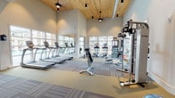 Located at 3 Woodcock Rd in Savannah, Georgia, this single-floor fitness center is a comprehensively outfitted facility designed to support a wide range of workout routines. The main workout area impresses with soaring vaulted ceilings finished in warm natural wood, lending the space an open, airy ambiance. Patterned carpet tiles define the workout zones, while large windows along the perimeter flood the interior with natural light and offer views of the surrounding community. The cardio section features a row of treadmills positioned directly in front of oversized windows, alongside elliptical trainers and stair climbers. A dedicated strength and functional training zone houses a multi-station cable machine, an adjustable weight bench, a full dumbbell rack, TRX suspension trainers, battle ropes, medicine balls, BOSU balance trainers, and exercise balls. A wall-mounted flat-screen TV and a built-in mini-fridge add convenience to the strength area. A separate cycling studio with hardwood-style flooring accommodates multiple stationary spin bikes, complemented by a mirrored wall, a large display screen for guided workouts, and bright windows overlooking the outdoors. Together, these spaces create a modern, well-lit fitness environment suited for a full spectrum of training needs.