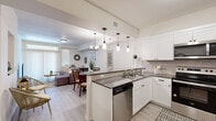 This single-floor residence offers a thoughtfully designed open-concept layout that seamlessly connects the kitchen, dining, and living areas. The kitchen is a standout feature, boasting crisp white cabinetry, dark countertops, a stylish tile backsplash, stainless steel appliances — including a side-by-side refrigerator, dishwasher, and microwave — and a peninsula with pendant lighting that doubles as a casual dining counter. The adjacent dining area is defined by an elegant chandelier and light-toned hardwood-style flooring that flows throughout the main living spaces. The living room is bright and welcoming, with large sliding glass doors and transom windows that invite natural light and provide outdoor views. The bedrooms are carpeted for comfort, featuring windows with sheer curtains that maintain a soft, airy feel. One bedroom is accompanied by a generously sized walk-in closet with built-in shelving and hanging rods, offering ample storage. The bathroom is cleanly finished with light wood-style flooring, white cabinetry, and a full tub-shower combination. Throughout, the space maintains a cohesive, modern aesthetic with neutral tones, clean lines, and warm natural accents.