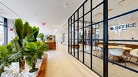 Located at 95 Anderson St in Hackensack, NJ, this single-floor commercial space offers a thoughtfully designed environment that balances open collaboration with private workspaces. The main area features a bright, open layout with large floor-to-ceiling windows that flood the interior with natural light and provide views of the surrounding streetscape. The polished light-toned tile flooring and crisp white ceilings with recessed and track lighting give the space a clean, contemporary feel throughout.

A curved amber leather sofa anchors the central lounge area, complemented by a rounded upholstered ottoman and globe pendant lights — creating an inviting and stylish gathering zone. A warm walnut wood reception desk with brass detailing serves as a welcoming focal point near the entrance, surrounded by lush tropical plants that add a natural, organic touch.

Sleek black steel-framed glass partition walls enclose multiple private offices and a conference room, maintaining visual openness while providing acoustic separation. The enclosed conference room features a round wooden table and modern white chairs beneath a circular pendant light. Additional private booths with rich green upholstered seating and textured accent walls offer semi-private meeting or lounge options. A built-in kitchenette with warm wood cabinetry rounds out the functional amenities of this well-appointed space.