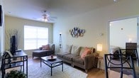 This single-floor space offers a comfortable and stylishly appointed living environment designed for both relaxation and everyday function. The living room serves as the heart of the space, centered around a plush, neutral-toned sofa accented with warm coral throw pillows, paired with a cushioned chaise lounge chair for additional seating. A soft, thick area rug anchors the seating arrangement around a sleek black metal coffee table, adding a touch of contemporary contrast to the room's warm palette. A large window fitted with white shutters allows abundant natural light to fill the room, creating a bright and airy atmosphere throughout the day. The ceiling features a fan with integrated lighting, ensuring year-round comfort and practicality. Decorative wall art — a cluster of colorful, textured circular pieces — adds a creative focal point and personal character to the space. Ambient floor lamps placed thoughtfully around the room provide warm supplemental lighting for evenings. The flooring throughout is hardwood, offering a clean and classic foundation that ties the entire space together. Overall, the space strikes a balanced blend of functionality and tasteful design, making it an inviting and livable environment.