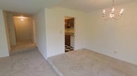This single-floor space offers a straightforward, functional layout with a clean and understated design. The main living area is finished with wall-to-wall neutral carpet, providing a soft, quiet foundation that contributes to the room's simple, uncluttered feel. White walls throughout keep the space bright and versatile, ready to accommodate a range of furnishings and décor styles. A wall-mounted candelabra-style light fixture adds a subtle decorative touch to the living area.

A doorway opening from the main living area leads directly into the kitchen, which features a striking black-and-white checkered tile floor that adds a classic, timeless character. The kitchen is equipped with dark wood cabinetry offering ample storage, a sink set into a countertop along the back wall, and a white dishwasher. The cabinetry includes glass-front upper cabinets, adding visual interest to the compact space. A hallway visible on the left side of the main area suggests additional rooms or storage beyond the main living space. Overall, the layout is practical and compact, making efficient use of the available floor plan.