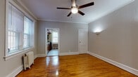 This single-floor unit offers a thoughtfully laid-out interior defined by warm, polished hardwood floors that extend throughout the living and sleeping areas. Each room is finished with crown molding and equipped with a ceiling fan, contributing to a classic yet comfortable ambiance. Neutral gray walls provide a clean, versatile backdrop across all spaces.

The living areas are generously proportioned, with multiple rooms offering ample space for various uses. Large windows dressed with blinds invite abundant natural light, while radiator heating units underscore the building's traditional character. One room opens to the outdoors through a set of glass-paned French doors, providing a seamless connection to the outside. A dedicated entry door adds convenience and privacy.

The kitchen is well-appointed with warm honey-toned wood cabinetry, granite-style countertops, and a full suite of black appliances including a gas range, built-in microwave, and dishwasher. A secondary door in the kitchen provides additional exterior access. Light ceramic tile flooring completes the space.

The bathroom features a full tub-and-shower combination enclosed by classic white subway tile, maintaining a clean and functional aesthetic. Throughout the unit, the combination of traditional architectural details and practical finishes creates a cohesive and inviting living environment.