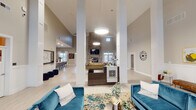The Greenview clubhouse is a thoughtfully designed, single-story community gathering space characterized by dramatically high ceilings and an open, airy floor plan. Light hardwood-style flooring runs throughout, complementing the neutral gray walls and bright white architectural columns that anchor the central common area.

The main lounge serves as the heart of the space, furnished with teal velvet sofas and accent chairs arranged around a patterned area rug. A floor-to-ceiling stacked stone fireplace wall, topped by a wall-mounted television, creates a striking focal point. Pendant lighting and recessed fixtures illuminate the area evenly.

Adjacent to the lounge, a casual recreation zone features a full shuffleboard table and additional seating. A well-appointed kitchenette area includes white cabinetry with under-cabinet lighting, a countertop bar with gray upholstered stools, and ample prep space suited for community gatherings.

A private conference room, accessible through double French doors, provides a dedicated space for meetings, outfitted with a central table and chairs. Additional seating nooks with teal armchairs, framed artwork, and decorative greenery are distributed throughout, offering a variety of comfortable settings. Natural light filters in through multiple windows and glass entry doors, enhancing the welcoming atmosphere of the overall space.