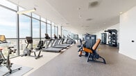 Located in the heart of downtown Saint Petersburg, FL, this high-rise fitness and wellness facility offers a comprehensive, resort-style experience across a single expansive floor. The centerpiece is a generously sized fitness center lined with floor-to-ceiling windows that frame sweeping panoramic views of the city and surrounding waterways, flooding the space with natural light. The gym is outfitted with a full suite of professional-grade equipment, including multiple treadmills, ellipticals, rowing machines, stair climbers, and an extensive selection of strength and resistance machines with distinctive tan leather accents. Rubberized flooring throughout the workout area ensures both comfort and durability. Adjacent to the gym, a dedicated spa area provides a tranquil retreat featuring private treatment rooms finished with herringbone wood-look tile flooring, built-in cabinetry with sink stations, and calming ocean-themed artwork. An indoor pool room clad in neutral large-format tiles offers a serene aquatic escape. A light-filled relaxation lounge, furnished with woven chaise lounges and framed by floor-to-ceiling glass, connects to an outdoor terrace with additional city views, completing this well-rounded wellness destination.
