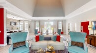 Located at 5003 Winster Dr in Winston-Salem, NC, this single-floor space offers a thoughtfully designed layout comprising a living room, kitchen, and hallway. The living room is the centerpiece of the space, boasting soaring vaulted ceilings that lend an open, airy feel. Large floor-to-ceiling windows and glass-paneled doors flood the area with abundant natural light while offering outdoor views of a pool and surrounding greenery. The living room is organized into distinct seating zones, featuring teal wingback chairs, a cream sofa accented with bold red and blue throw pillows, and plush area rugs over light hardwood flooring. A mounted flat-screen TV and globe pendant lighting add functional elegance to one lounging nook. Vibrant accent walls in red and teal punctuate the neutral gray-and-white palette, creating a dynamic, contemporary atmosphere. The kitchen is equally striking, featuring dark espresso cabinetry topped with crisp white countertops and a large central island with seating. A wood-paneled accent wall adds warmth and texture, complemented by decorative gear-shaped mirrors and globe pendant lights. A built-in coffee station and compact refrigerator enhance the space's utility. The hallway connects these areas seamlessly, contributing to an overall layout that is both functional and visually cohesive.