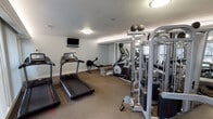 This fitness center is a compact yet fully outfitted facility designed to support a complete workout routine. The space is laid out on a single level with carpeted flooring and neutral-toned walls that create a clean, focused environment. Flush-mount ceiling lights provide consistent illumination throughout, while a row of large windows fitted with vertical blinds along one wall invites natural light and offers views of the surrounding greenery, allowing users to exercise in a bright and pleasant atmosphere. A full-length mirrored wall runs along one side of the room, effectively expanding the visual space and allowing users to monitor their form during workouts. The cardio section is well-represented with multiple treadmills, an elliptical trainer, and a rowing machine, catering to a variety of aerobic exercise preferences. A wall-mounted flat-screen television is positioned for easy viewing from the cardio area. The strength training zone features a multi-station cable machine with adjustable pulleys and a weight stack, accompanied by a workout bench, supporting a wide range of resistance exercises. A wall-mounted AED device and informational postings reflect a commitment to safety. The room is accessed through a classic French door, adding a refined architectural touch to the functional space.