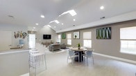 This single-floor space offers a well-designed, open-plan layout that blends comfort and functionality. The vaulted ceiling is a standout architectural feature, fitted with multiple skylights that allow abundant natural light to pour in, complementing the recessed lighting throughout. Light-toned luxury vinyl flooring runs across the entirety of the space, enhancing the bright and airy atmosphere.

The room is organized into distinct zones. A casual lounge area is anchored by upholstered gray sofas arranged on a textured area rug, with a wall-mounted flat-screen television serving as a focal point. Adjacent cabinetry provides practical storage. Nearby, a round dining table with modern gray chairs offers an intimate setting for small gatherings. A counter-height bar area along one wall includes sleek white stools, ideal for casual seating or individual use.

Neutral taupe walls are accented by colorful wall art with green and organic motifs, adding visual interest. Multiple windows fitted with roller shades allow for adjustable natural light. The overall ambiance is clean, contemporary, and welcoming, suited for communal or social use.