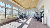 Located in Spring, TX, this fitness center offers a thoughtfully designed exercise space across a single floor. The room's architectural centerpiece is a bold yellow tray ceiling accented by recessed lighting and dual ceiling fans, creating a bright and energizing atmosphere overhead. Large, floor-to-ceiling windows line multiple walls, flooding the space with natural light and providing open views of the surrounding community and outdoor areas. The flooring features durable, patterned carpet tile suited for high-traffic fitness use. The space is comprehensively equipped with a diverse array of cardio machines, including treadmills, ellipticals, and a stationary cycling bike, arranged to take full advantage of the natural light from the windows. A multi-station cable strength training machine and a weight bench with a rack provide ample options for resistance training. A dumbbell rack along one wall ensures free weights are readily accessible. Full-length mirrors on one wall visually expand the space and support proper form during workouts. A wall-mounted flat-screen television serves as an entertainment focal point. Glass double doors provide access to an outdoor area, enhancing ventilation and connectivity to the broader facility.