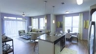 This thoughtfully designed single-floor space showcases a seamless open-concept layout that connects the kitchen, dining, and living areas into one cohesive and inviting environment. The kitchen serves as the centerpiece, anchored by a large island with a dark countertop, undermount sink, and pendant lighting that adds a modern, polished touch. Dark cabinetry contrasts elegantly with the lighter finishes throughout the space. Rich hardwood flooring runs continuously across the entire floor plan, grounding the design with warmth and texture. The living area is generously proportioned, featuring a comfortable seating arrangement, a ceiling fan, and large windows dressed with patterned curtains that allow abundant natural light to fill the room. The lavender-toned accent walls create a bold yet sophisticated color palette, complemented by pops of green and yellow in the décor and soft furnishings. A dedicated dining area sits adjacent to the kitchen, offering an intimate setting for meals. Multiple windows throughout the space ensure bright, airy conditions and provide views to the surrounding exterior. The overall ambiance is modern and vibrant, blending functional design with a distinctive, stylish aesthetic.