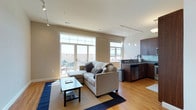 This single-floor open-plan space combines a living area and kitchen in a clean, contemporary layout. The living area is anchored by a light grey sectional sofa paired with a sleek rectangular coffee table atop a layered blue and neutral rug. Large floor-to-ceiling windows and a glass-paned door flood the space with natural light while offering views of the surrounding neighborhood and direct access to a private outdoor patio, where seating is visible. Track lighting runs across the ceiling, supplementing natural light throughout the day. Warm-toned hardwood flooring extends seamlessly from the living area into the kitchen, reinforcing the open, cohesive feel of the space. The kitchen features rich dark wood cabinetry, granite-look countertops, a teal glass tile backsplash, and stainless steel appliances including a dishwasher and sink. Pendant lights hang above the kitchen workspace, adding focused illumination and a modern touch. The neutral wall tones and clean lines throughout contribute to an airy, well-organized ambiance suited to everyday living and entertaining.