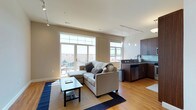 This single-floor open-plan space combines a living area and kitchen in a clean, contemporary layout. The living area is anchored by a light grey sectional sofa paired with a sleek rectangular coffee table atop a layered blue and neutral rug. Large floor-to-ceiling windows and a glass-paned door flood the space with natural light while offering views of the surrounding neighborhood and direct access to a private outdoor patio, where seating is visible. Track lighting runs across the ceiling, supplementing natural light throughout the day. Warm-toned hardwood flooring extends seamlessly from the living area into the kitchen, reinforcing the open, cohesive feel of the space. The kitchen features rich dark wood cabinetry, granite-look countertops, a teal glass tile backsplash, and stainless steel appliances including a dishwasher and sink. Pendant lights hang above the kitchen workspace, adding focused illumination and a modern touch. The neutral wall tones and clean lines throughout contribute to an airy, well-organized ambiance suited to everyday living and entertaining.