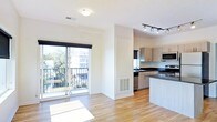 This single-floor space features an open-concept layout that seamlessly connects the living area and kitchen. Wide-plank hardwood flooring runs throughout, lending warmth and continuity to the interior. A large sliding glass door floods the living area with natural light and opens onto a private balcony, offering views of a tree-lined neighborhood streetscape. A central structural column with a built-in HVAC vent adds architectural interest while dividing the space subtly without enclosing it.

The kitchen is well-appointed with dark countertops that contrast against light cabinetry, providing ample storage and prep space. A kitchen island extends the counter area and offers additional seating or workspace. Stainless steel appliances, including a built-in microwave and dishwasher, are integrated into the design. Track lighting above the kitchen provides focused, adjustable illumination, complementing the natural light from a dedicated kitchen window. The overall ambiance is clean, contemporary, and functional, with neutral tones and modern fixtures creating a cohesive aesthetic throughout the space.