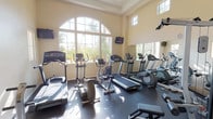This fitness center is thoughtfully designed to support a comprehensive workout experience within a single-story facility. The space is characterized by soaring ceilings that create an open, airy atmosphere, enhanced by an impressive arched window wall that floods the room with natural light and offers views of surrounding palm trees and outdoor amenities. Clerestory windows positioned along the upper walls provide additional daylight throughout the interior.

The cardio zone is well-appointed with multiple treadmills, elliptical trainers, upright and recumbent stationary bikes, and a rowing machine, all arranged to take advantage of the natural light and views. Wall-mounted flat-screen televisions are positioned for convenient viewing during workouts. A full-length mirror spans one wall, amplifying the sense of space and allowing users to monitor their form.

The strength training area includes a cable machine, weight benches, and free weights, providing options for a full-body workout. The dark rubberized flooring throughout is both durable and functional. A glass-paned French door provides entry to the space, and the warm, neutral-toned walls contribute to the clean, welcoming ambiance of this well-maintained fitness facility.