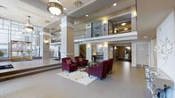 This grand lobby is defined by its impressive double-height ceiling, which creates an expansive, airy atmosphere immediately upon entry. A decorative mezzanine level overlooks the main floor, framed by ornamental iron railings and supported by classic white columns that add architectural distinction throughout the space. A sweeping staircase connects the two levels, serving as both a functional and visual centerpiece.

The main floor features large-format light grey tiles that extend throughout, lending a clean and polished appearance. A central seating arrangement consists of rich burgundy upholstered armchairs positioned around a low coffee table atop a patterned area rug, providing an inviting gathering space. Recessed lighting and wall sconces offer warm, layered illumination complemented by a prominent pendant light fixture near the entrance.

Neutral taupe and white wall tones provide a refined backdrop, while framed artwork contributes subtle visual interest. A reception or service area is visible in the background, along with additional corridors suggesting access to further amenities. The overall ambiance is polished, professional, and welcoming.